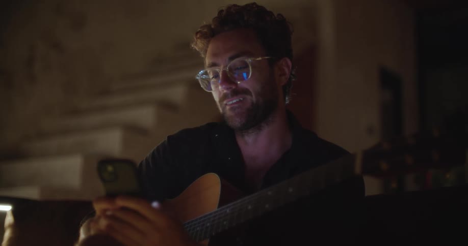 A man with curly hair and glasses texting on a phone while holding a guitar