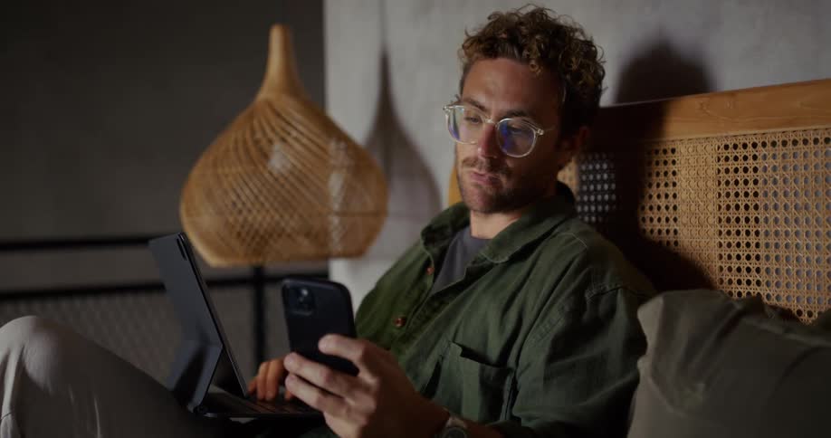 A man relaxes on a couch using a smartphone and tablet with notebooks nearby suggesting casual work or study