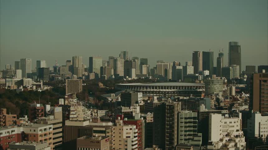 A wide shot captures the tokyo cityscape with numerous buildings under a hazy sky with the japan national stadium visible in the midground