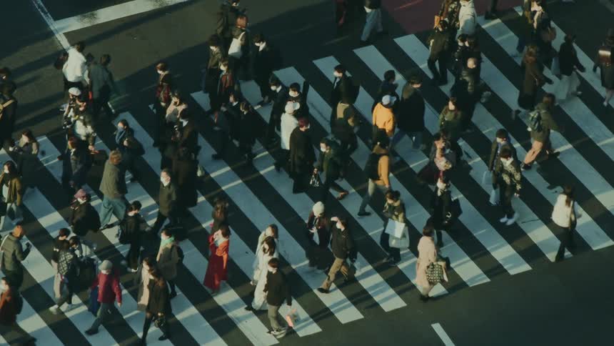 A large crowd of pedestrians crosses a city intersection at a busy crosswalk seen from an aerial perspective