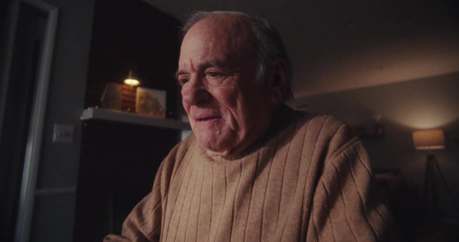 An elderly man is sitting indoors using a laptop and smiling warmly in a softly lit cozy room