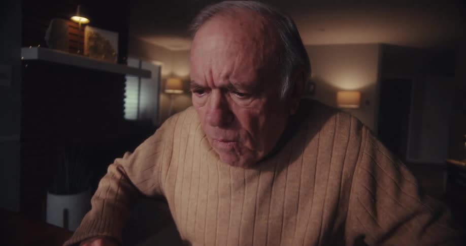 An elderly man sits indoors at a desk focused and slightly confused while looking at a laptop screen