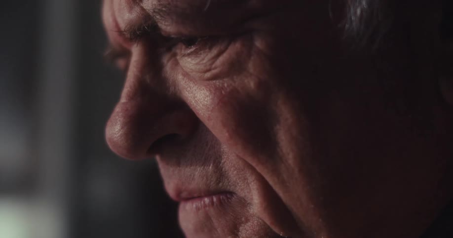 Closeup of an elderly mans face in a dimly lit room showing alternating expressions of happiness and deep thought