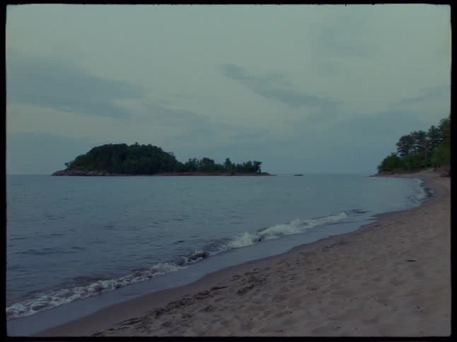 A calm empty beach at dusk with gentle waves and a distant treecovered shoreline under a cloudy sky