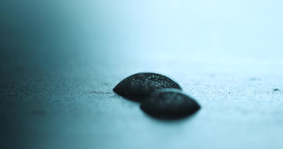 Two dark dome shaped semi spheres with a bubbling texture sit in sharp focus on a blue lit grainy surface resembling abstract droplets