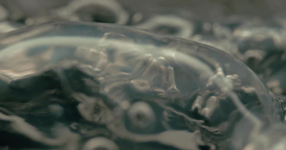 Transparent bubbles slowly form and pop over eerie metallic humanoid shapes submerged beneath a glossy surface