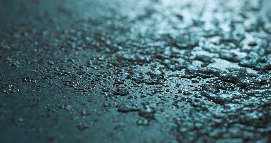 An extreme close up of a wet textured surface possibly asphalt shows glistening water droplets under cool blue lighting