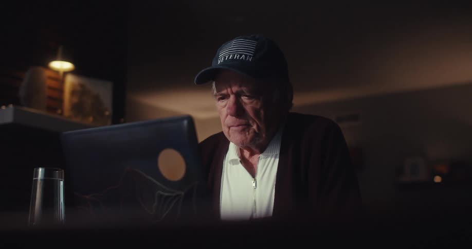 Elderly man wearing a cap looks at a laptop screen in a softly lit indoor environment