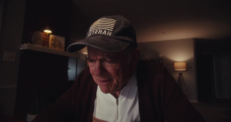 Elderly man sitting at a table in the living room looking at a laptop and laughing
