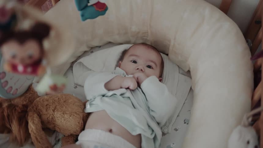 A baby lies in a crib with a mobile overhead with plush toys hanging from its frame