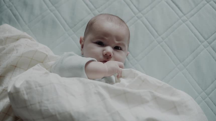 A baby is lying on a quilted mattress partially covered by a white blanket with a subtle checkered pattern