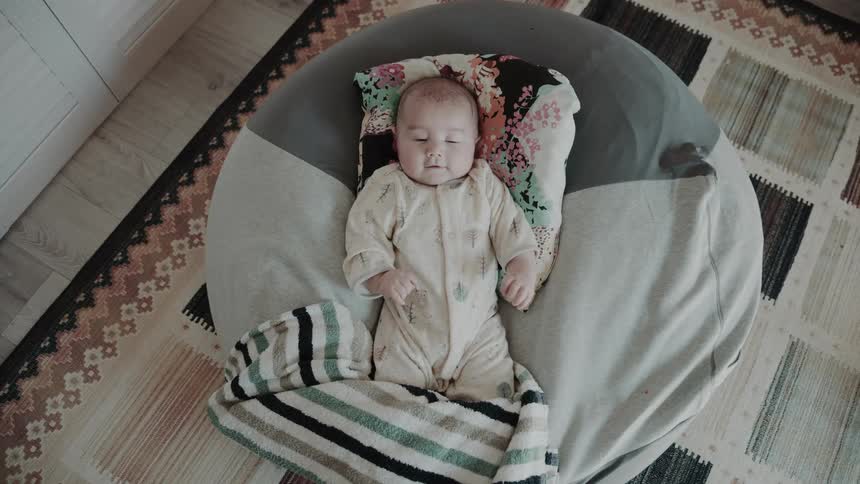 A baby is lying on a round cushion with a colorful pillow under the head and a striped blanket partially covering the body surrounded by a patterned rug and wooden floor