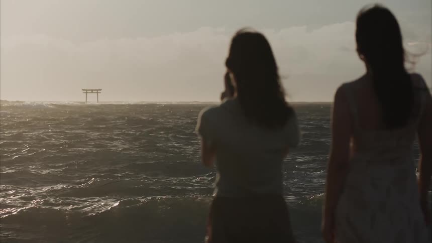 Two people stand on the shore one capturing the rough sea and distant torii gate on the horizon using a phone