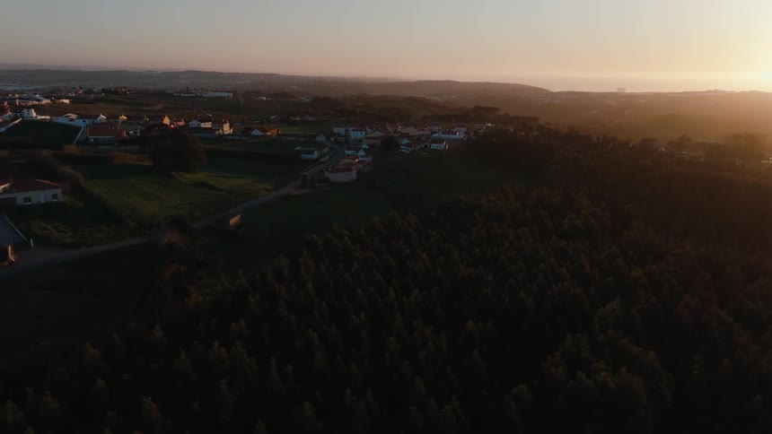 Sun shines from the horizon onto a rural town bordering a conifer forest on a foggy day
