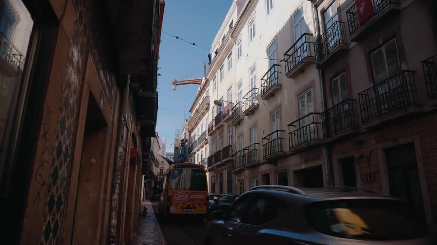 A narrow lisbon street features parked cars along tram tracks flanked by buildings with wroughtiron balconies under a clear sky