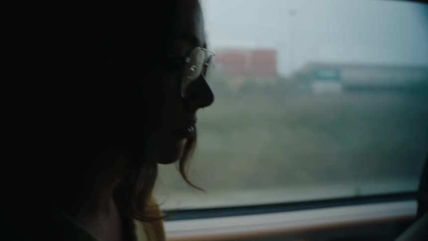 A close up of a woman with glasses and long hair wearing a yellow sweater engrossed in a book while seated on a train with shipping containers visible through the dimly lit window during daytime