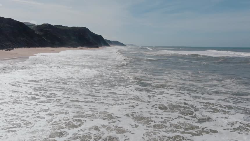 A vast expanse of choppy whitecapped waves crashes onto a sandy beach at the foot of dark rocky cliffs under a cloudy sky