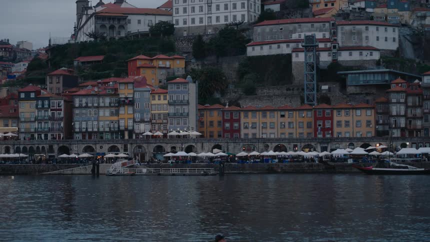 People walk along on both sides of the river passing a mountainside town on a cloudy day