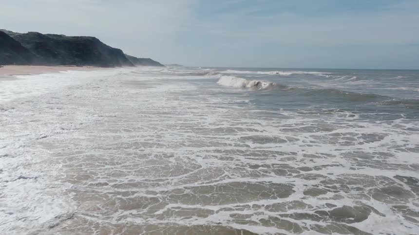 A vast expanse of choppy whitecapped waves crashes onto a sandy beach at the foot of dark rocky cliffs under a cloudy sky