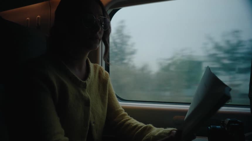 A woman is sitting on a train reading a book with a camera placed on the table next to the window showing a view of trees and buildings outside