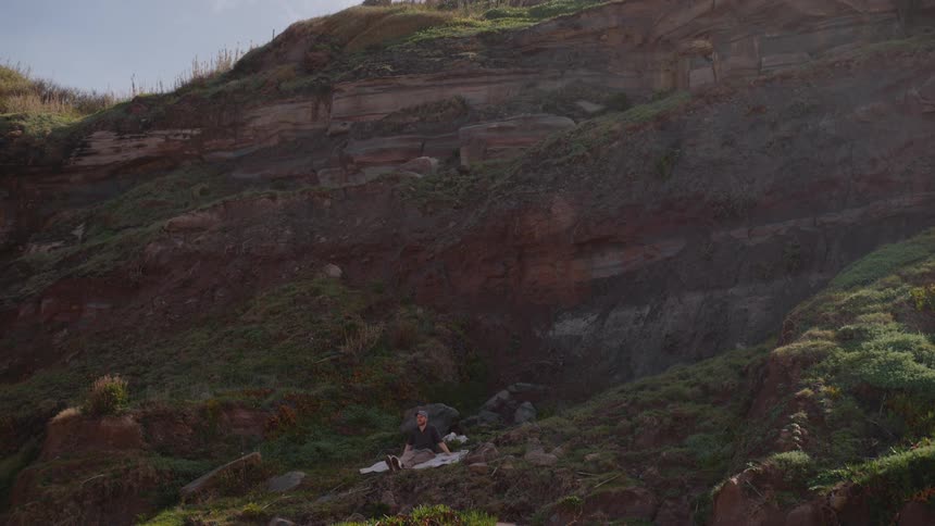 A lone individual sits crosslegged on a lightcolored cloth nestled amongst the rugged vegetationdotted terrain at the base of a rocky slope