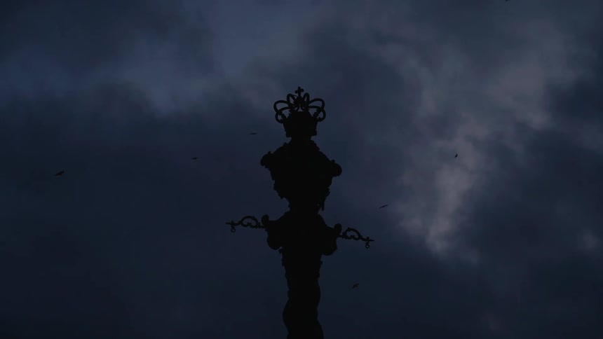 Christian sculpture with a crown and cross under a cloudy sky filled with flying birds