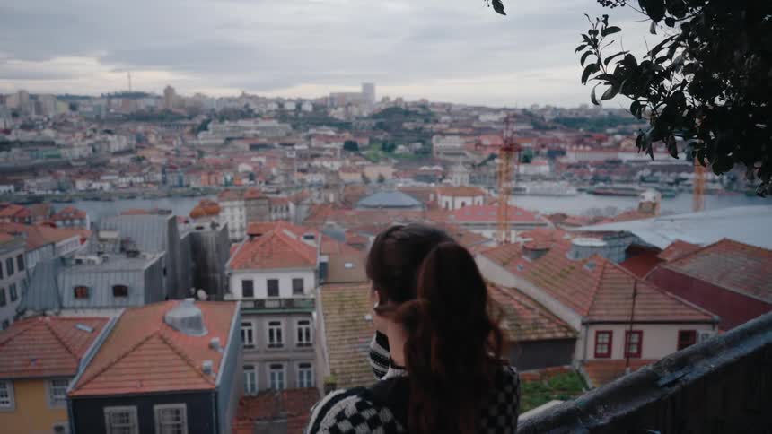 A woman with long hair stands next to a tree on an elevated platform gazing at the city skyline with numerous residential buildings visible in the midday light