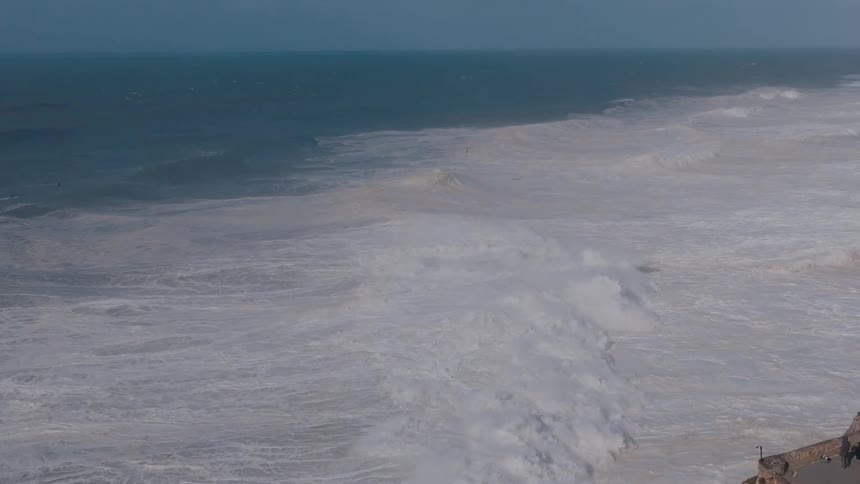 Two people stand on a ledge overlooking the coast as waves roll in to shore and seagulls fly over the water