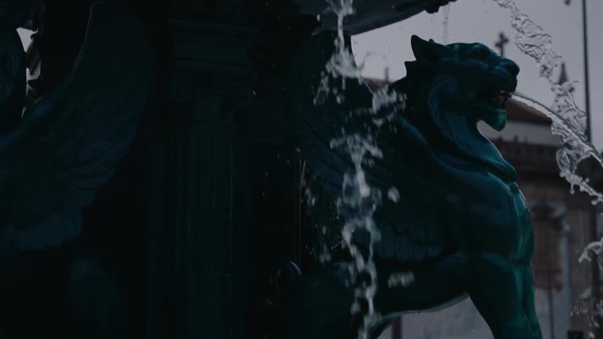 A dark moody shot of a fountain with water flowing from sculpted spouts and lion figures in an urban setting
