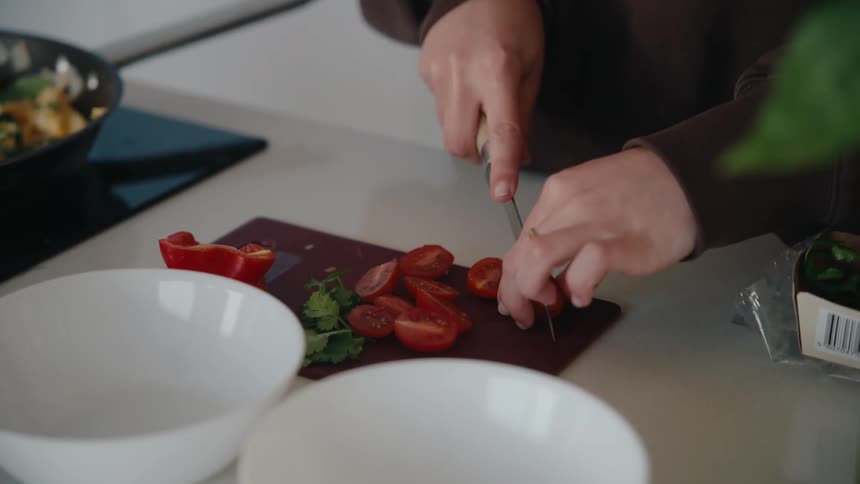 A person wearing a ring cuts cherry tomatoes on a dark cutting board surrounded by bowls and other food items on a light countertop
