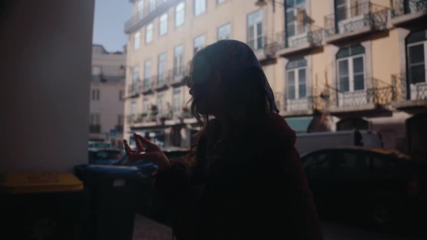 A woman stands in shadow holding a cigar on a narrow city street with buildings and parked cars behind her