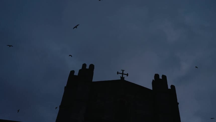 A church building with a cross on top stands silhouetted against the dusk sky as birds fly overhead
