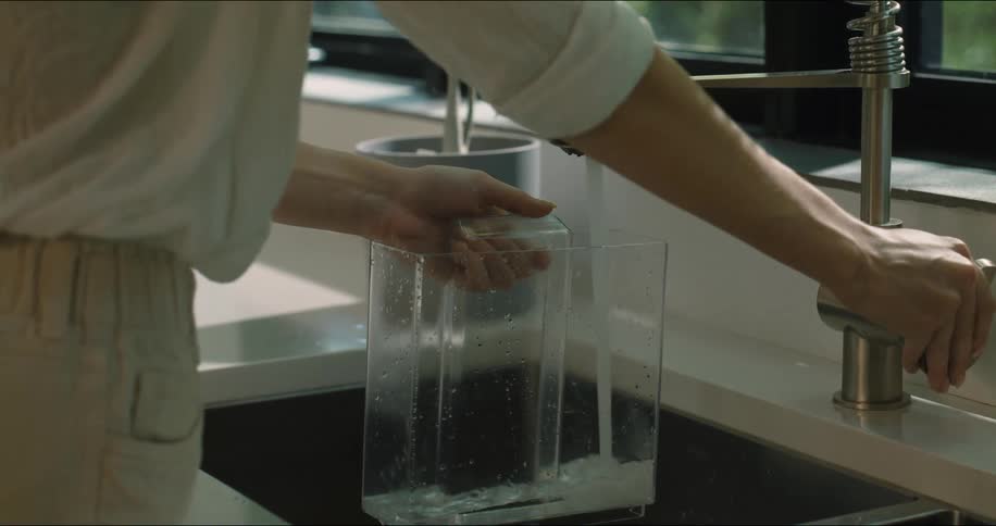 A person fills a glass jug with water from a kitchen sink tap