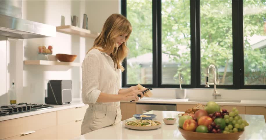 A woman stands in a modern kitchen preparing food on a counter with a bowl of assorted fruits nearby and large windows in the background