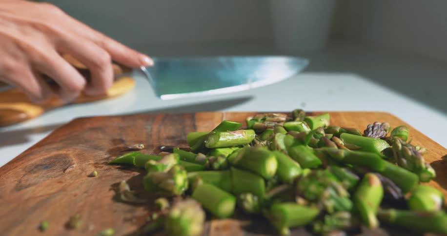 A close up of a hand chopping fresh green asparagus on a wooden cutting board in a brightly lit kitchen