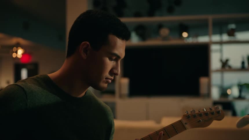 A young man plays electric guitar in a cozy home studio focused on his computer amid warm lighting and creative gear