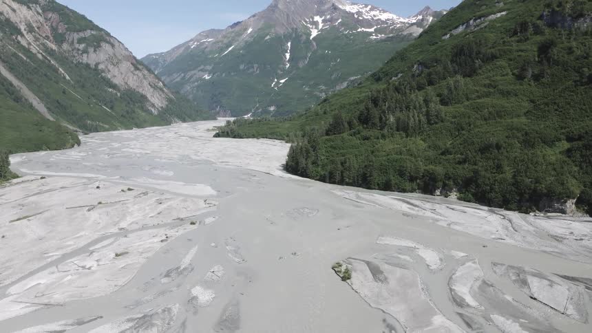 Aerial view of a wide dry riverbed surrounded by lush green mountains with patches of snow under a clear sky