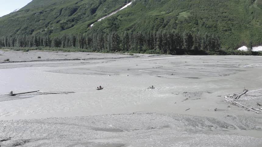 Aerial view of rafters navigating a braided glacial river through a remote mountain valley with dense forests and snow capped peaks