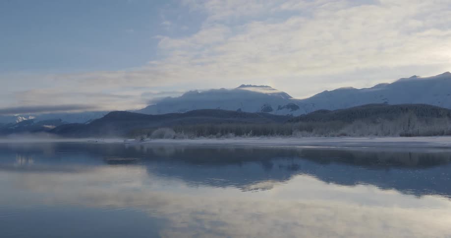 A wide shot shows a calm lake reflecting snowy mountains and a cloudy sky with mist rising in a peaceful setting