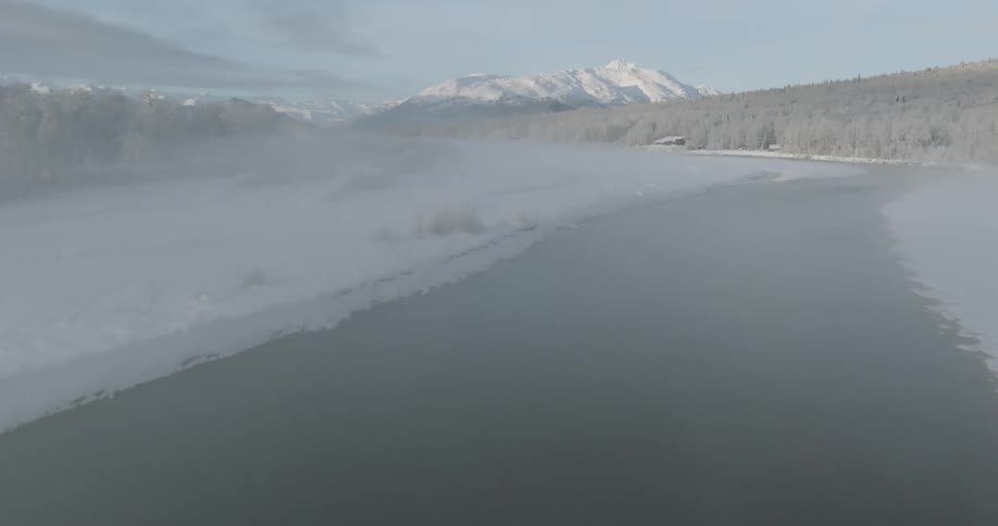 An aerial view follows a misty river flowing through a snowcovered landscape with frosted trees and distant mountains as birds fly over the icy terrain