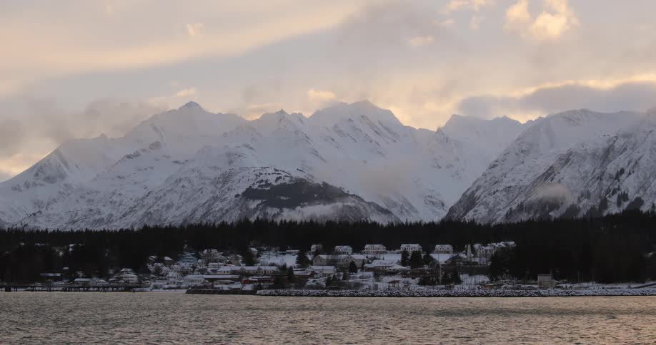 Snowcovered village sits on the waters edge with towering mountains in the background at dusk