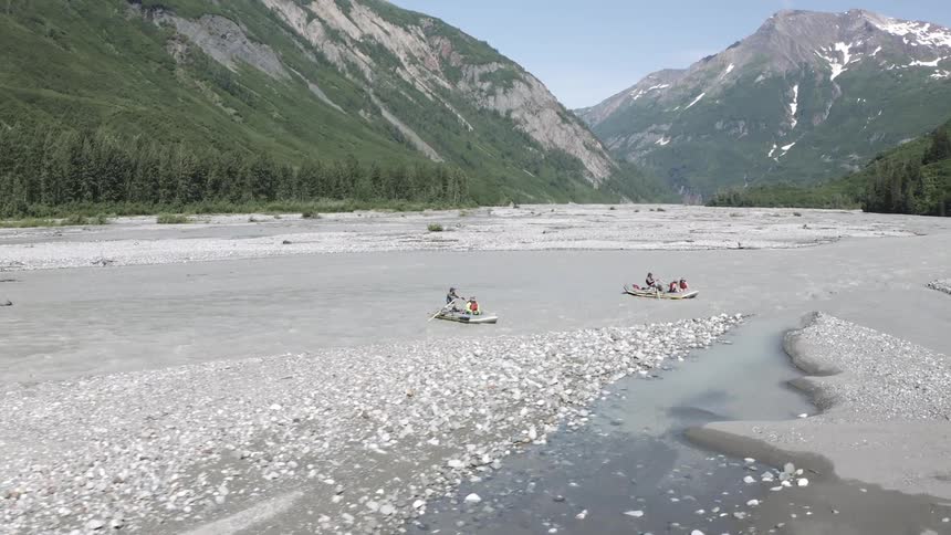 Two canoes each with two people rowing on a wide river surrounded by many trees and mountains in the background