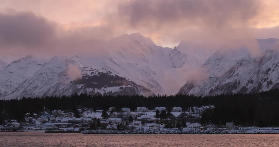 A wide shot captures a serene coastal town beneath snowcovered mountains at dawn or dusk with mist and calm water enhancing the tranquil setting