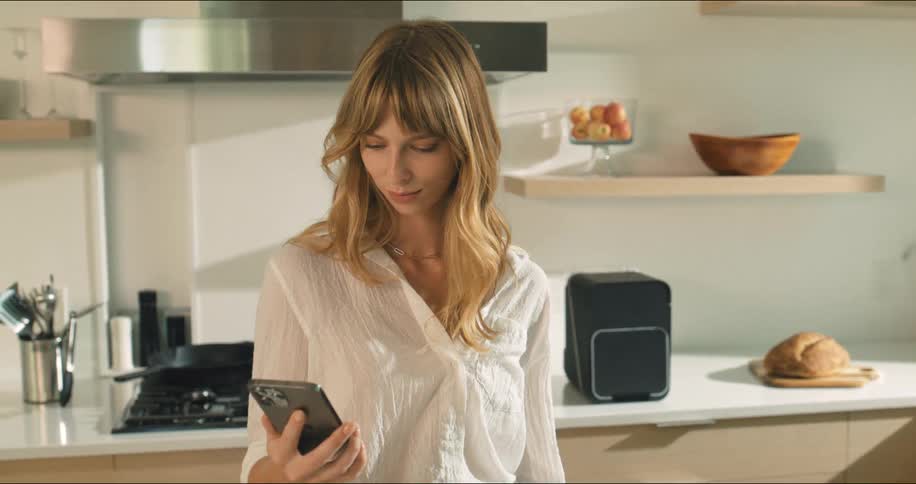 A woman stands in a sunlit modern kitchen looking at her smartphone while casually preparing for the day beside a countertop loaf of bread