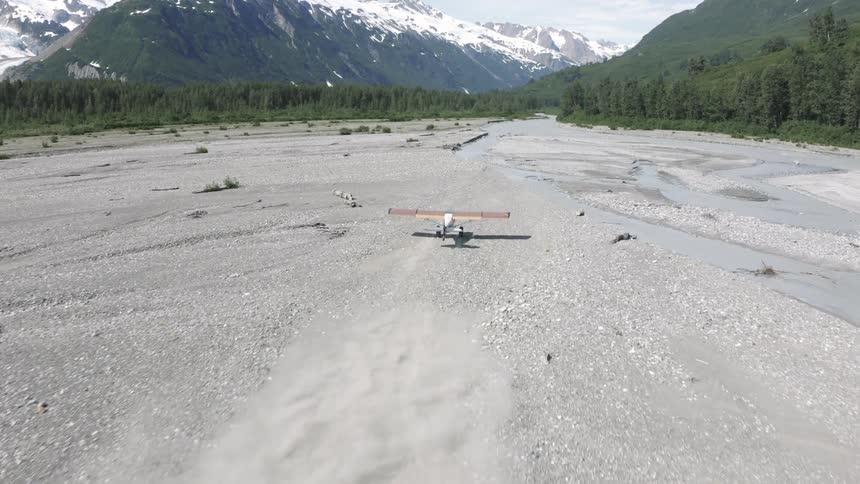 Small plane takes off from a dry riverbed surrounded by dense forest and snowy mountains in the background