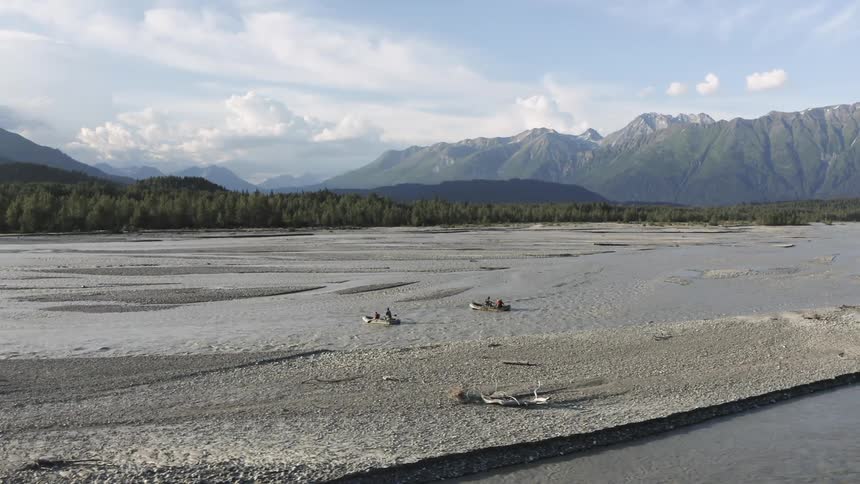 Several people paddle inflatable rafts down a wide shallow river flanked by gravel bars and forest with towering mountains in the background under a clear blue sky