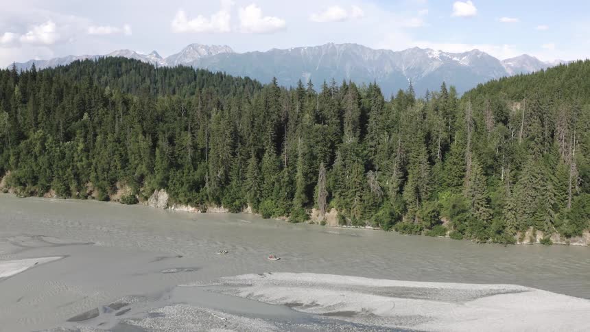Aerial view of a forested mountain valley with a winding river snowcapped peaks and small rafts on the water