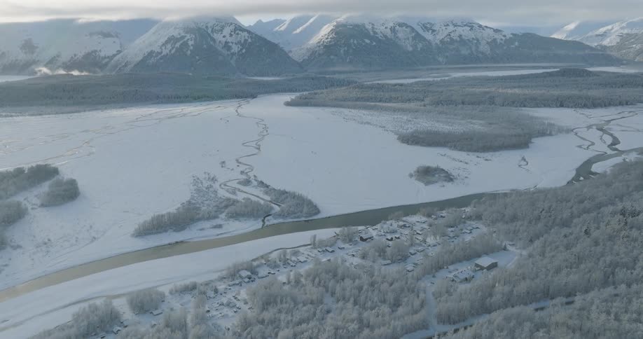 An aerial view of a small town blanketed in snow at midday with a frozen lake nearby and a snowcovered mountain visible in the distance