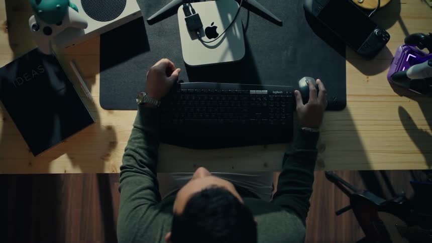 A young man sits intently focused at a desk in a dimly lit room working on a computer with multiple monitors and rgb lighting while the camera orbits around him
