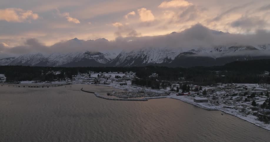An aerial view captures a serene coastal town nestled between a snowy mountain range and calm waters at dawn or dusk with a visible harbor and light snow covering the landscape